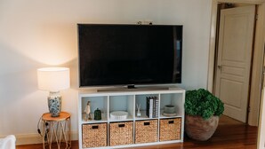 Un salón con una televisión de pantalla plana sobre un soporte blanco, una lámpara, cestas de mimbre y una planta en maceta.
