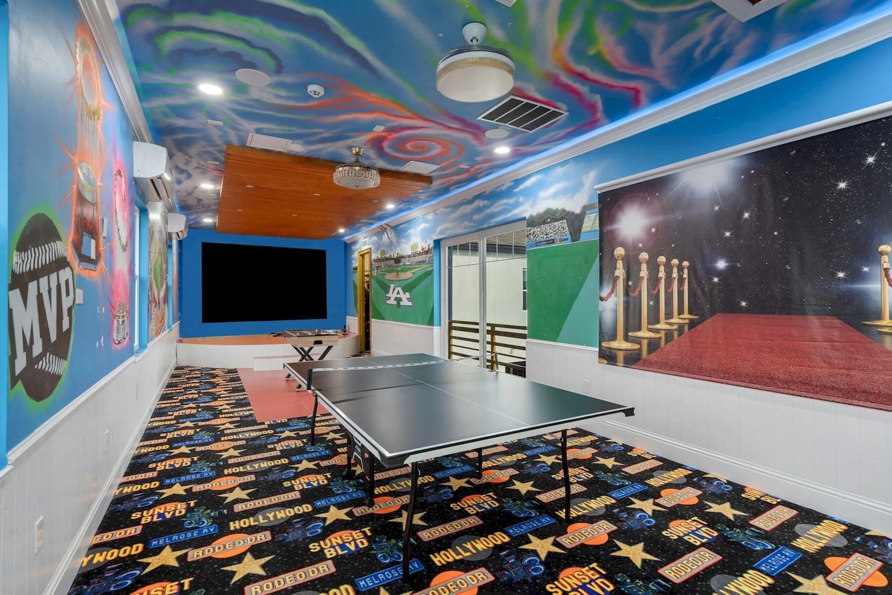 A room with a ping pong table, a large screen, and a mural of a baseball field.