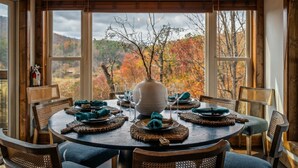 A dining area with a round table set for four, surrounded by wicker chairs. The table is adorned with a vase, plates, and glassware. Above the table, there are four hanging pendant lights. Large windows with a view of autumn foliage are visible. - Designer Cabin | Mtn Views | Game Room | Hot Tub (Sevierville)