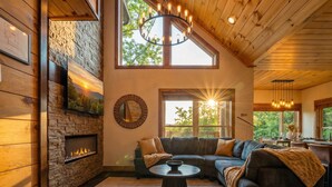 A cozy living room with a stone fireplace, a large window with a view of trees, and a modern chandelier. - Designer Cabin | Mtn Views | Game Room | Hot Tub (Sevierville)