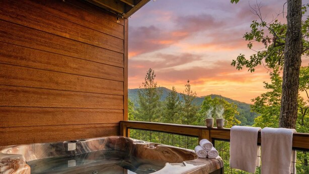 A wooden deck with a hot tub, towels, and a view of a sunset over a forested landscape. - Designer Cabin | Mtn Views | Game Room | Hot Tub (Sevierville)