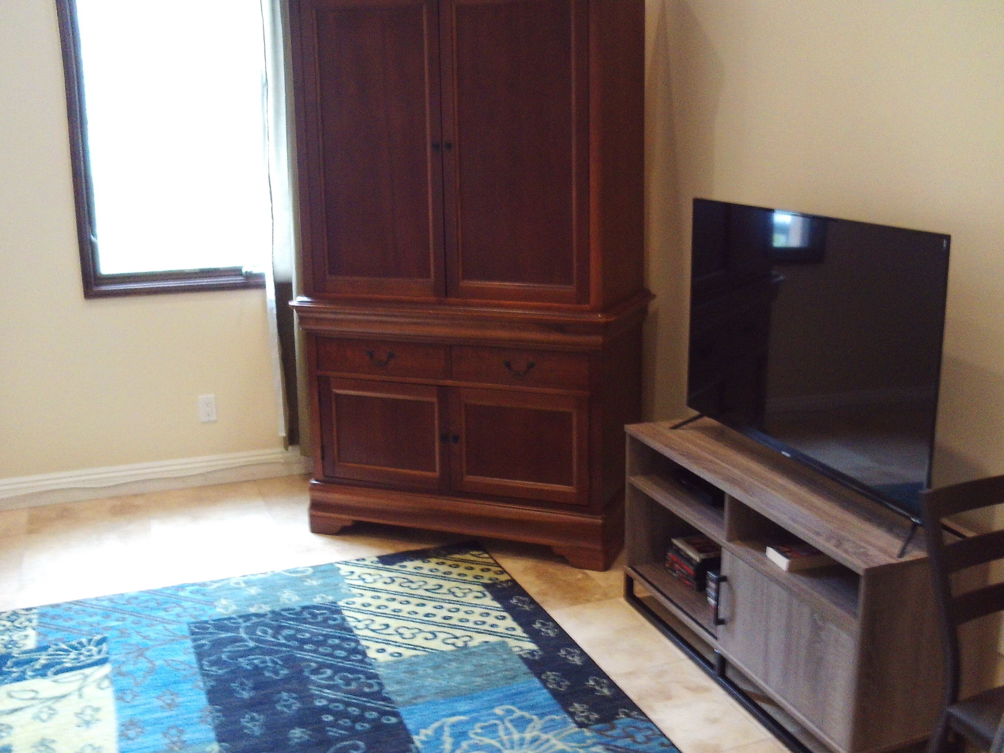 Ein Zimmer mit einem großen Holzschrank, einem Flachbildfernseher auf einem Standfuß und einem gemusterten Teppich.