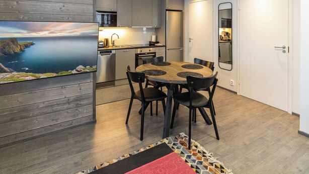 Une cuisine moderne dotée d’appareils électroménagers en acier inoxydable, un coin repas avec une table en bois et des chaises noires, et une vue sur la mer par une grande fenêtre.