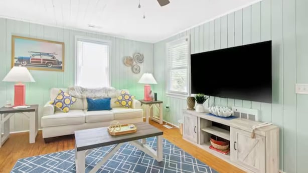 A living room with a ceiling fan, a flat-screen TV, a sofa with patterned cushions, a coffee table, and a cabinet with decorative items. - Last minute and Fall specials! Walk to beach! 4br 4ba Perfect location  (North Myrtle Beach)