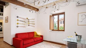 A red sofa, a glass table with wine glasses, a window, and wall art. - La Torre del Biscone   Campo dè Fiori (ROMA)