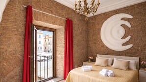 A hotel room with a brick wall, a large bed with striped bedding, a chandelier, and a view of buildings through a sliding door. - La Torre del Biscone   Campo dè Fiori (ROMA)