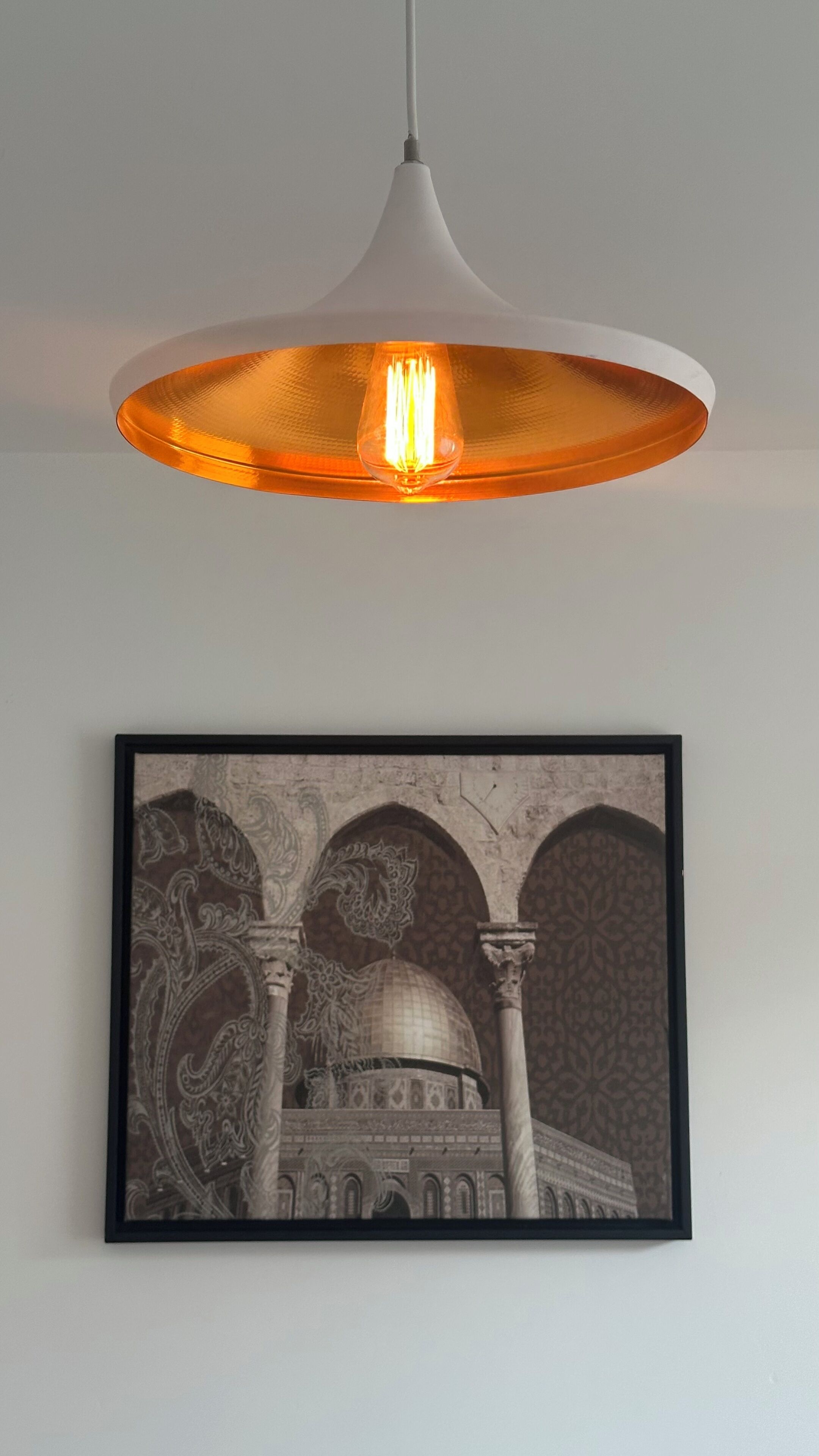Un luminaire suspendu, avec un abat-jour blanc et un intérieur orange, est accroché au-dessus d’une reproduction architecturale encadrée qui orne le mur.