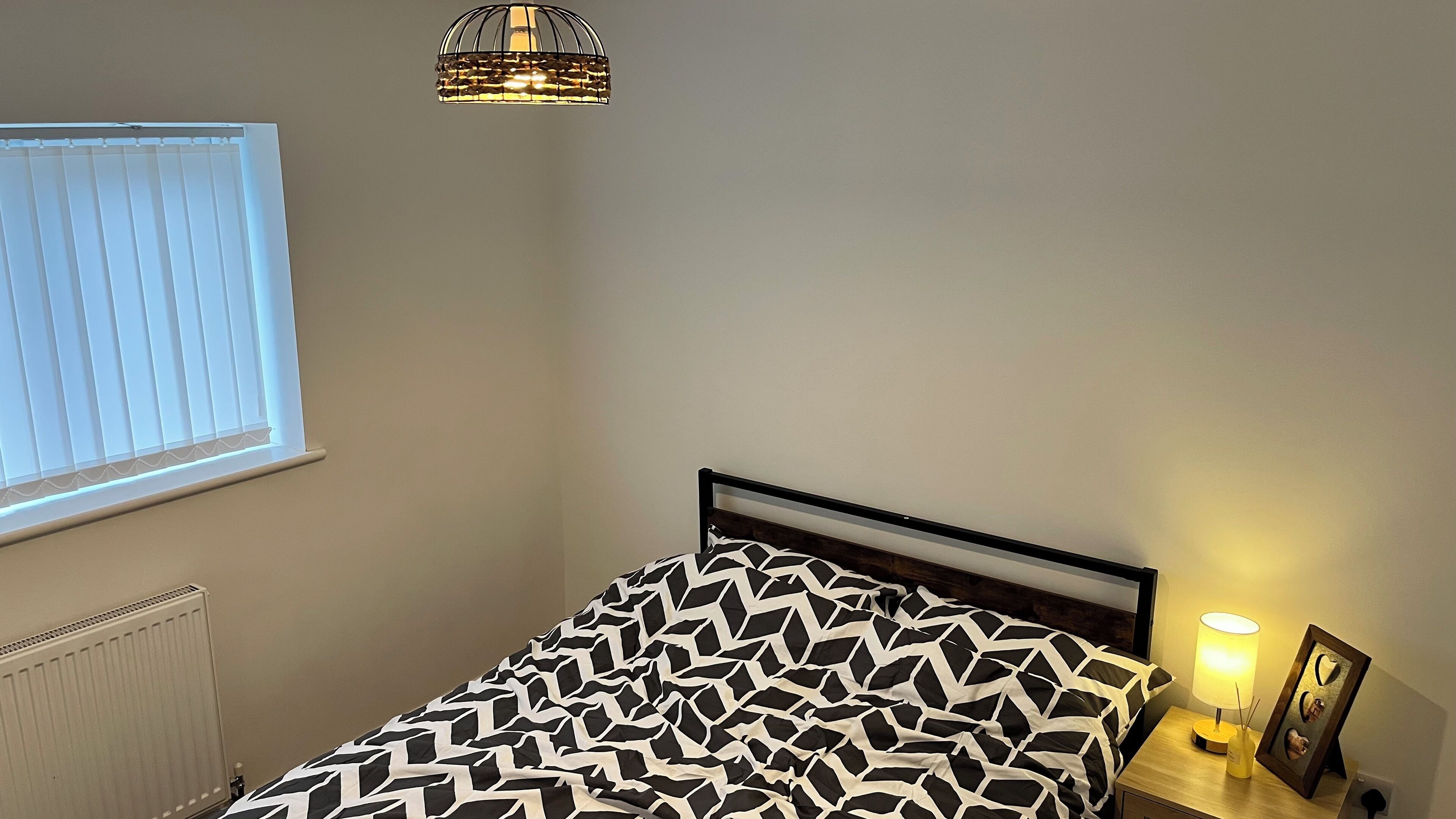 Een slaapkamer met een bed, nachtkastje met lamp, raam met zonwering en een radiator.
