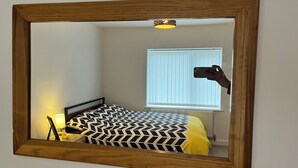 Un dormitorio con cama, lámpara de noche, ventana con persianas y espejo con marco de madera.