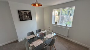 Un comedor con mesa de vidrio, cuatro sillas, una lámpara colgante, un cuadro en la pared y una ventana con vistas a la vegetación.