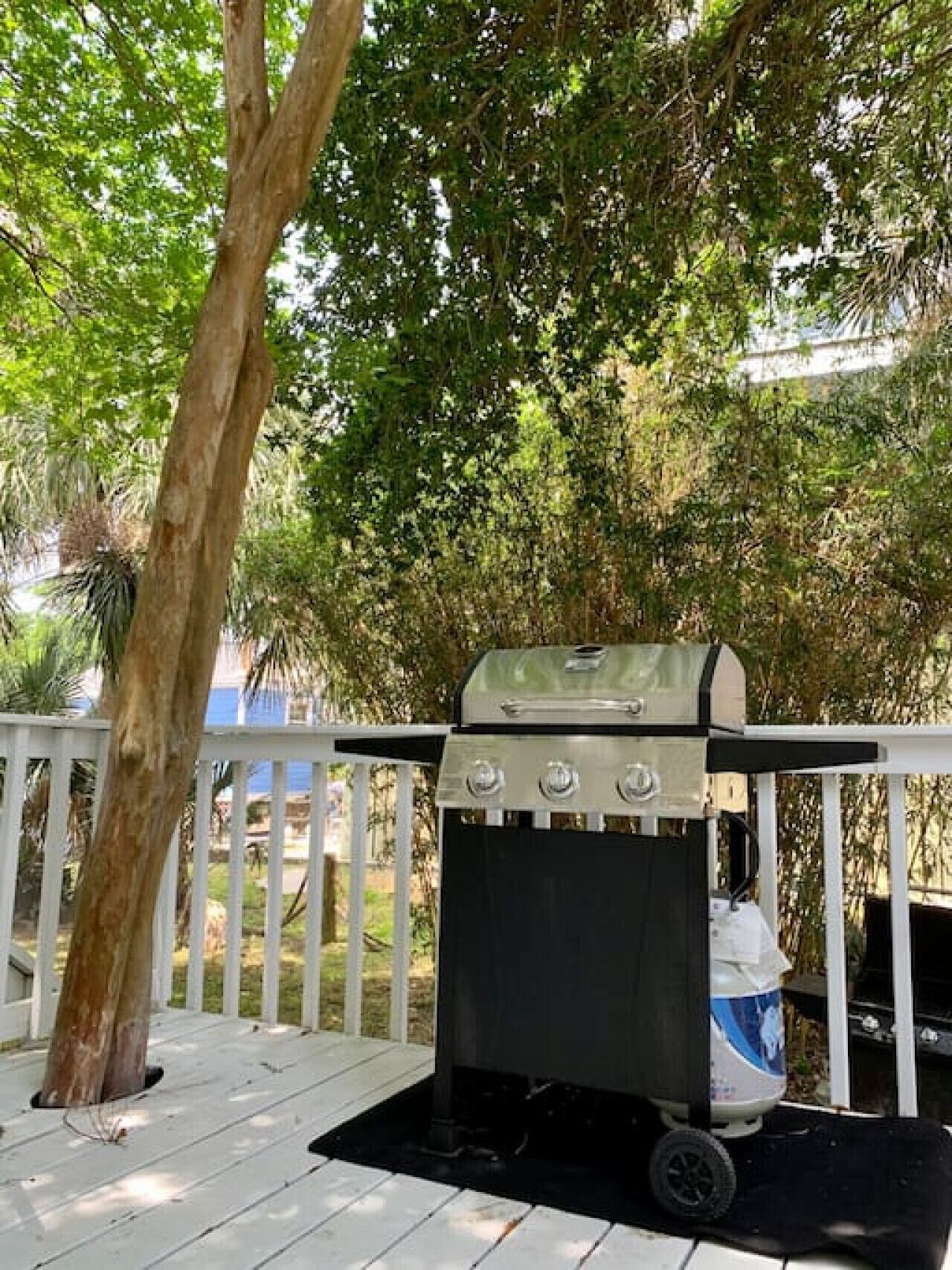 A portable grill on a wooden deck with a white railing, surrounded by lush greenery.