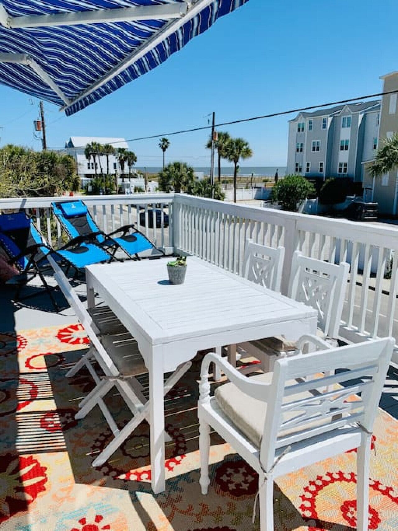 A white outdoor seating area with a table and chairs, a blue umbrella, and a patterned rug.