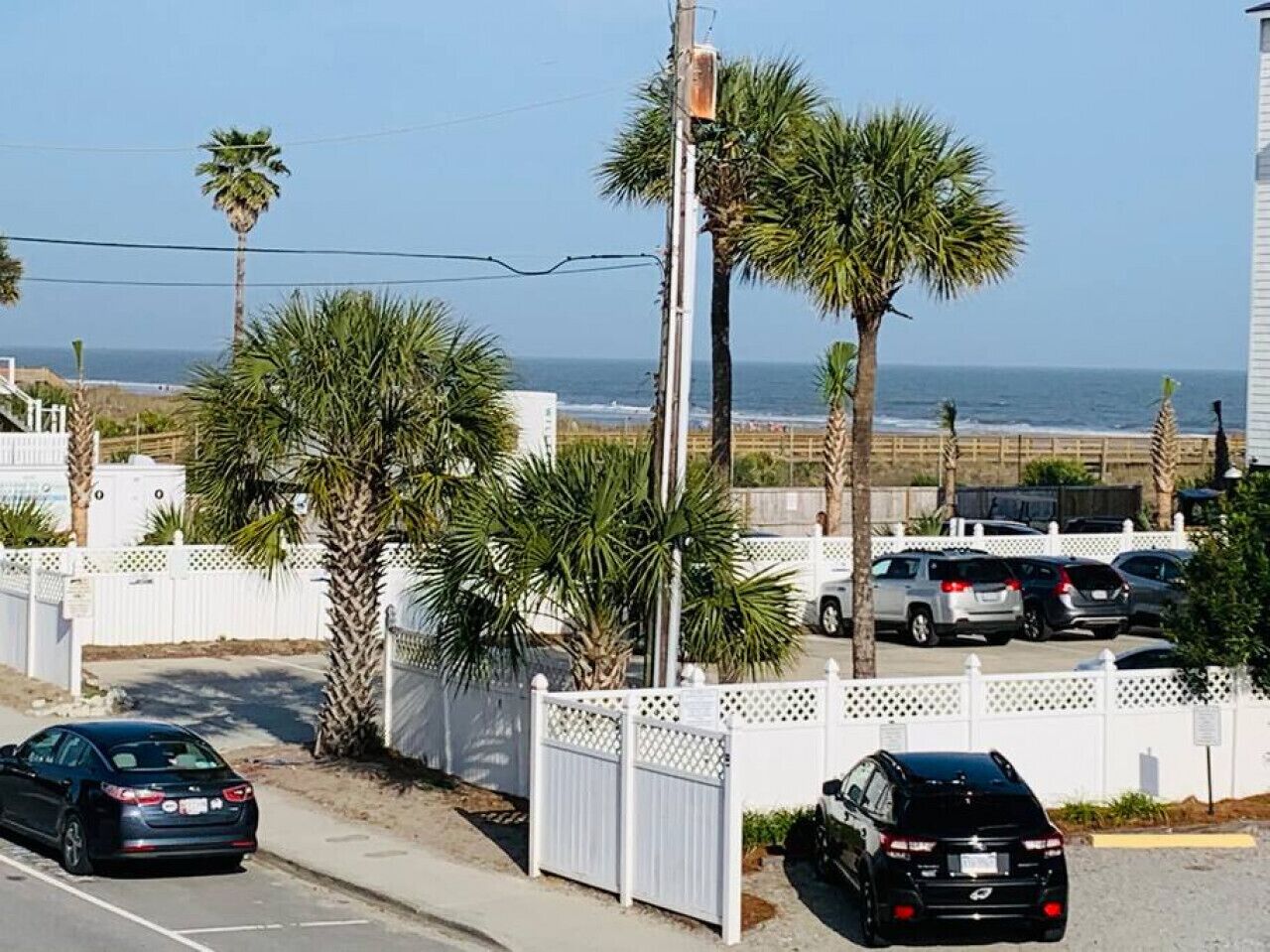 A residential street with parked cars, palm trees, and a white fence.