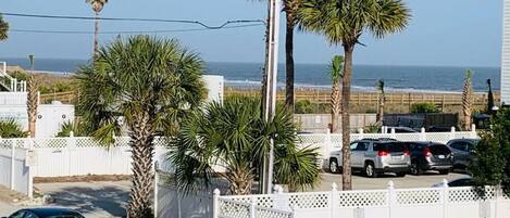 A residential street with parked cars, palm trees, and a white fence.