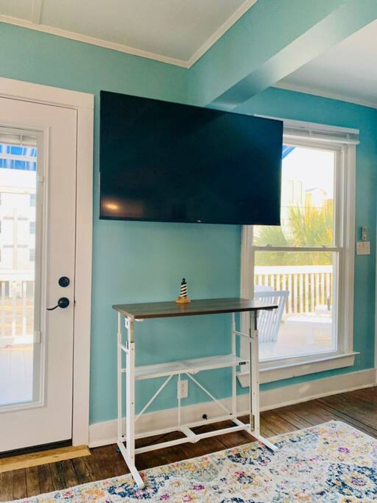 Une pièce aux murs de couleur turquoise, avec une porte blanche, un téléviseur fixé au mur, un bureau en métal blanc et un tapis à motifs floraux.