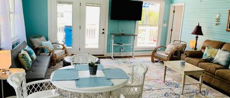 A living room with teal walls, a white ceiling fan, a TV mounted on the wall, a dining table with chairs, a sofa, and a patterned rug.