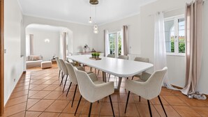 Un comedor amplio con una mesa larga y seis sillas, un sofá al fondo y vista a zonas verdes exteriores.