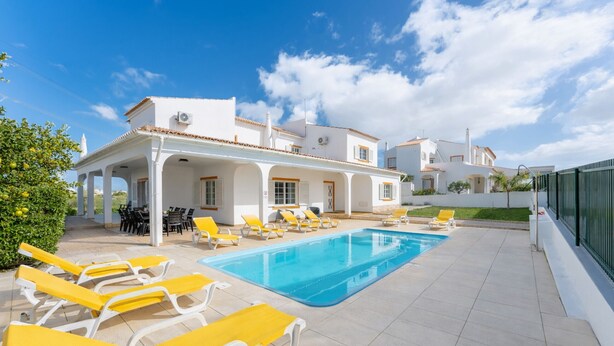 Un área de piscina con sillas de descanso amarillas, un espacio cubierto para sentarse y un edificio blanco con tejado de azulejos.