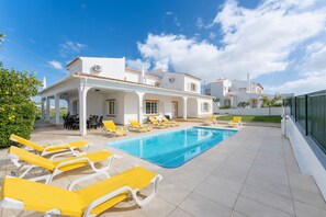 Un espace piscine avec des chaises longues jaunes, un coin salon abrité et un bâtiment blanc doté d’un toit en tuiles.