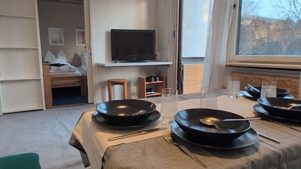 A dining area with a table set for four, a television mounted on the wall, and a window with a view of trees. - Vacation home with large terrace in Schöneiche near Berlin (Schöneiche bei Berlin)