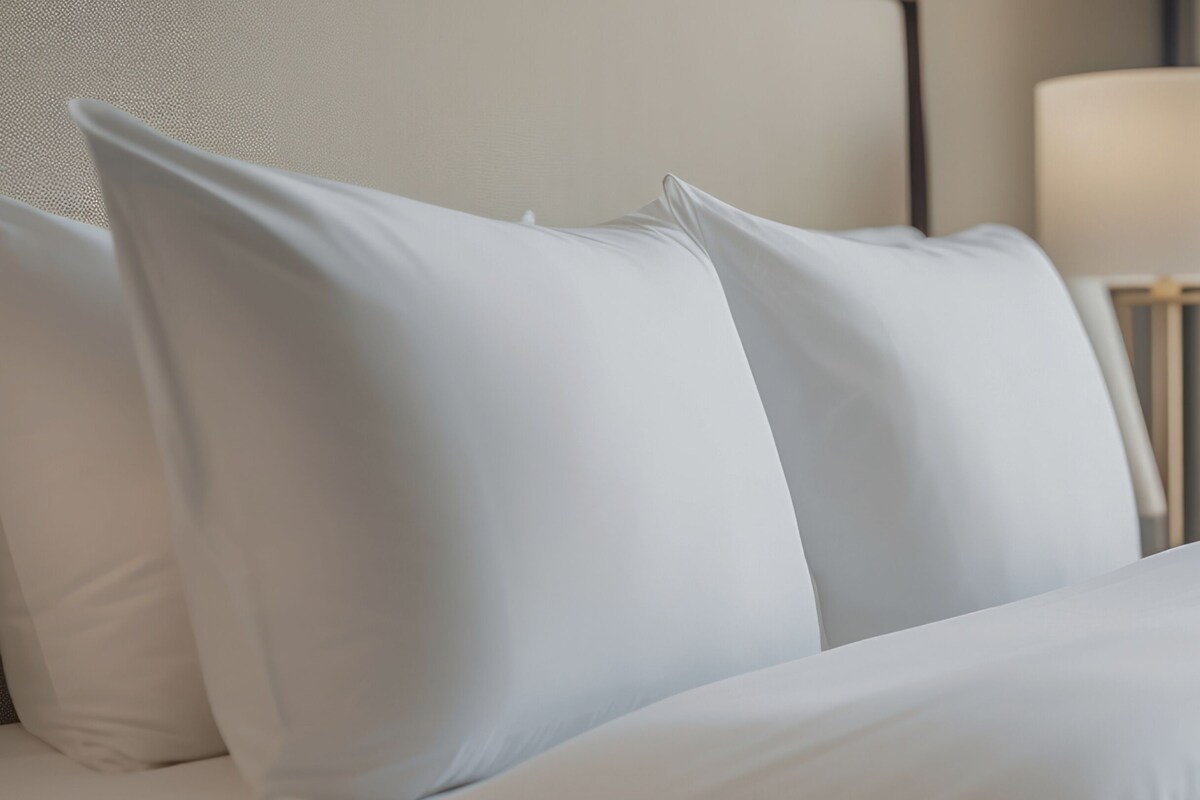 Una cama con ropa blanca y almohadas, una lámpara de noche y una pared con textura.