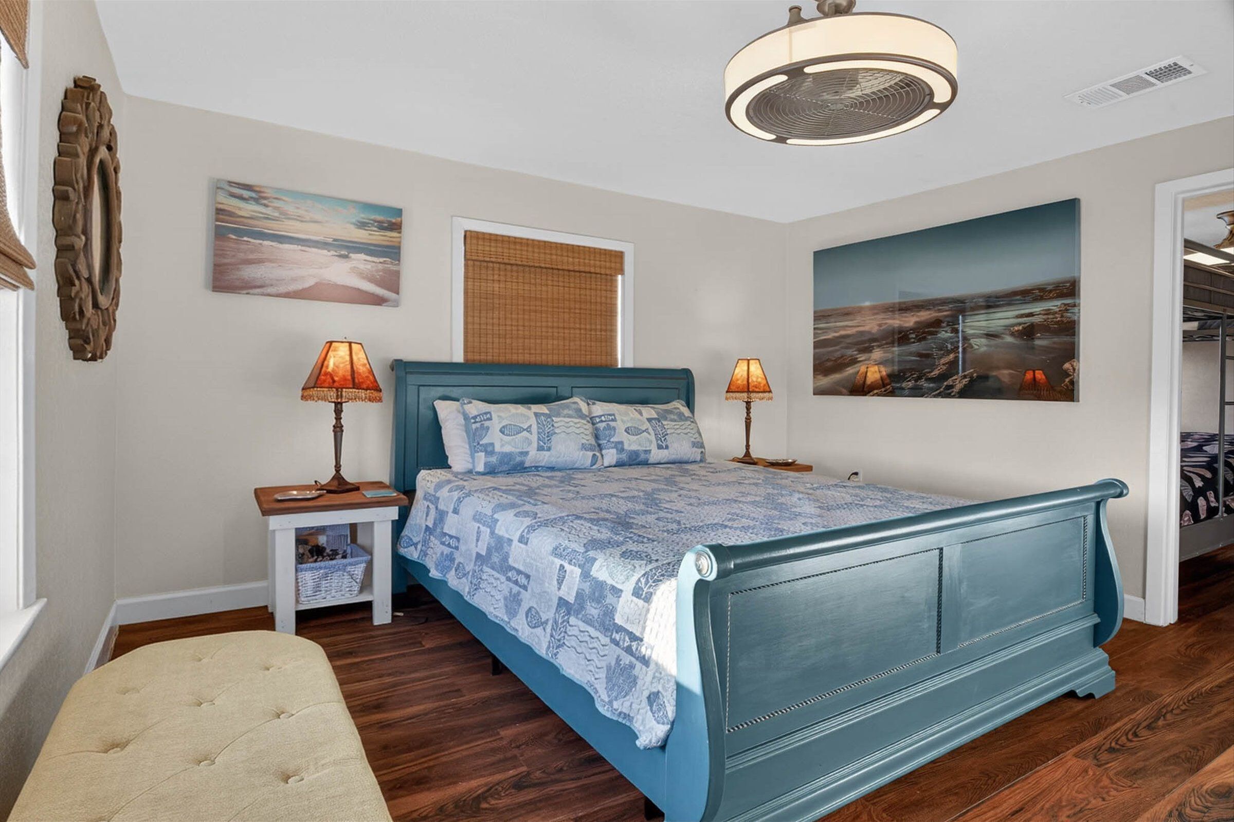 A bedroom with a blue bed, two bedside lamps, a wooden nightstand, a bench, a large window with blinds, and two wall-mounted paintings.