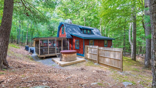 Une cabane dotée d’une structure en verre, d’un bain à remous et d’une terrasse en bois, située dans un cadre forestier.