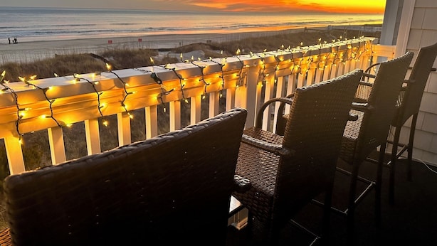 A balcony with wicker chairs, a railing adorned with string lights, and a view of the ocean at sunset. - Sea Cloisters 303a (North Myrtle Beach)