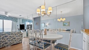 A dining area with a round table, white chairs, and a chandelier. There is a sofa with patterned upholstery and a large window with curtains. - Sea Cloisters 303a (North Myrtle Beach)