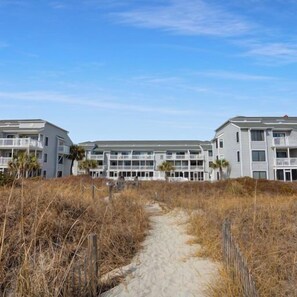 A beachfront hotel with a sandy pathway leading to the entrance, surrounded by dry grass and palm trees. - Sea Cloisters 303a (North Myrtle Beach)