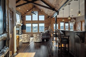 Interior de una cabaña de troncos con chimenea de piedra, vigas de madera en el techo y ventanas amplias que ofrecen vistas al exterior.