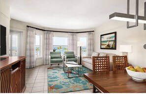 Uma sala de estar moderna com TV, poltronas verdes, um sofá branco, mesa de jantar de madeira e vista para o oceano.