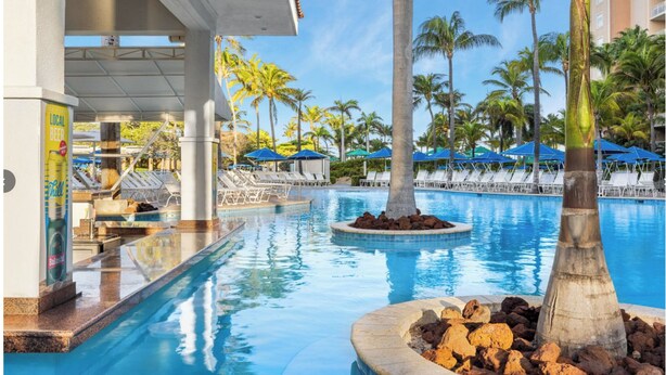A pool area with lounge chairs, palm trees, and a sign for local beer. - Ocean View/Ocean Side  2BR at Aruba Surf Club – Steps to Palm Beach Paradise (Noord)