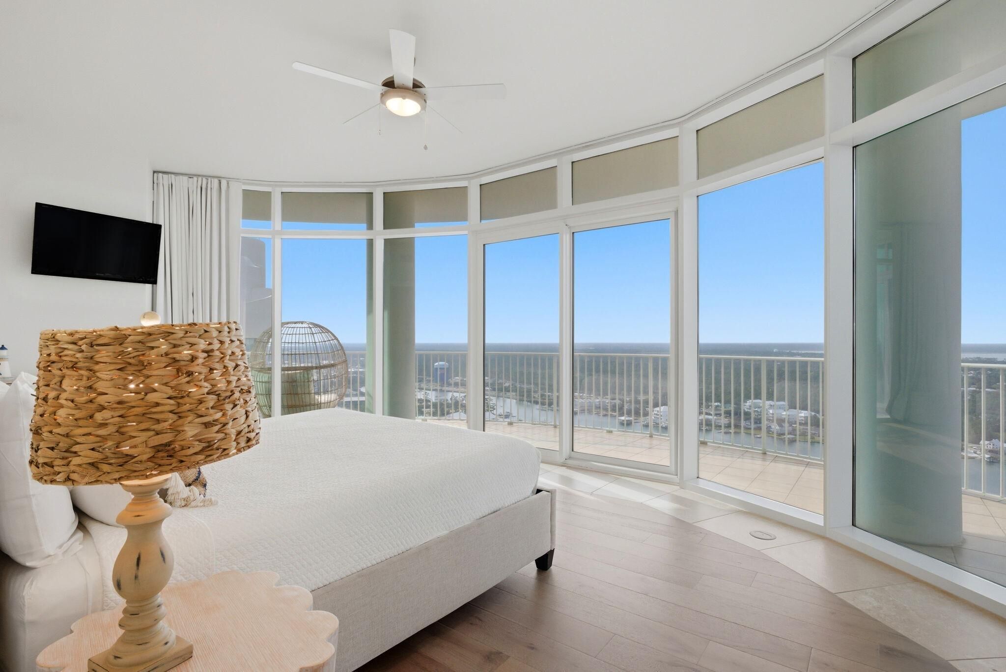 A bedroom with a large bed, a ceiling fan, a TV on the wall, and a balcony with a view of the city.
