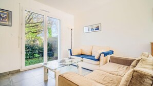 Uma sala de estar moderna com uma mesa de centro de vidro, sofás bege e uma porta de correr de vidro que dá para um jardim.