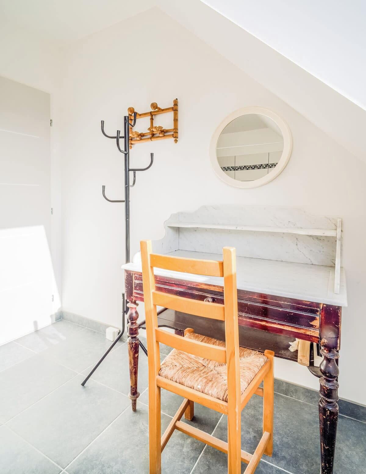A piano with a white and red finish, a wooden chair with a woven seat, a coat rack with a circular mirror, and a tiled floor.