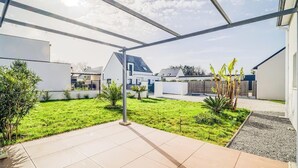 Um pátio moderno com piso de azulejos, rodeado por um gramado bem cuidado e diversas plantas.