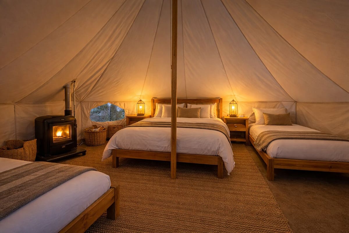 A tent with two beds, a wood-burning stove, and a view of trees through a window.