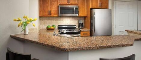 Una cocina con encimeras de granito, armarios de madera, microondas de acero inoxidable, refrigerador y cafetera.