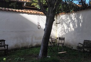 Un patio con un muro blanco, un árbol y una hamaca. Hay dos bancos de madera y una mesa con sillas.