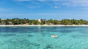 A beach with clear turquoise water, a person kayaking, and a building with a white facade and balconies overlooking the beach. - Luxury Beachfront 2BR | Private Beach, Pools, Rooftop | 10 Min from Tulum (Tankah Cuatro)
