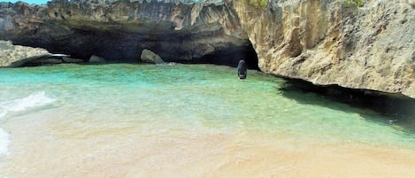 Una playa con agua turquesa cristalina, la entrada a una cueva rocosa y una persona cerca de la cueva.