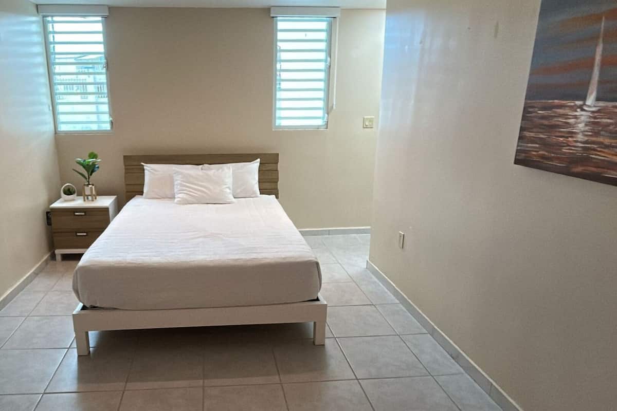 A bedroom with a bed, a nightstand, a potted plant, and a painting of sailboats on the wall.