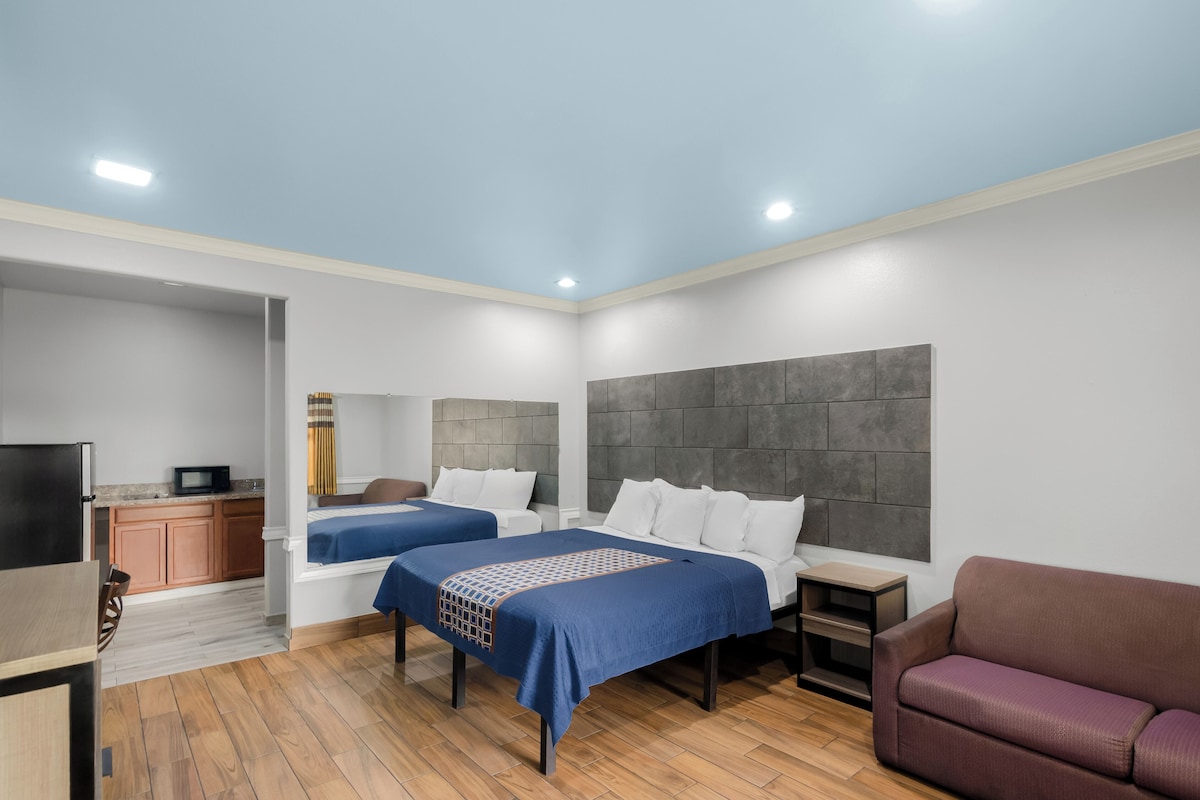 A modern hotel room with two beds, a TV, a small kitchenette, and a seating area.