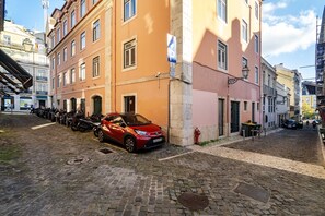 Una strada acciottolata con auto e motociclette parcheggiate, edifici con balconi e un cielo sereno.