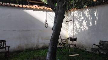 Un patio con una pared blanca, un árbol y un columpio. Hay dos bancos de madera y una mesa con sillas.
