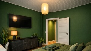 Ein Schlafzimmer mit grünen Wänden, einem Bett mit grüner Bettwäsche, einem an der Wand montierten Fernseher, einem Nachttisch mit Lampe und einer Tür zu einem anderen Raum.
