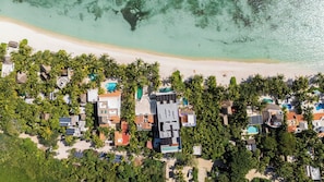 Vue aérienne d’une station balnéaire comprenant plusieurs bâtiments, une plage de sable et une mer turquoise aux eaux limpides.