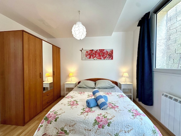 Une chambre à coucher comprenant un lit, des lampes de chevet, une armoire en bois, une couvre-lit à motifs floraux, une œuvre d’art murale et une fenêtre avec des rideaux.