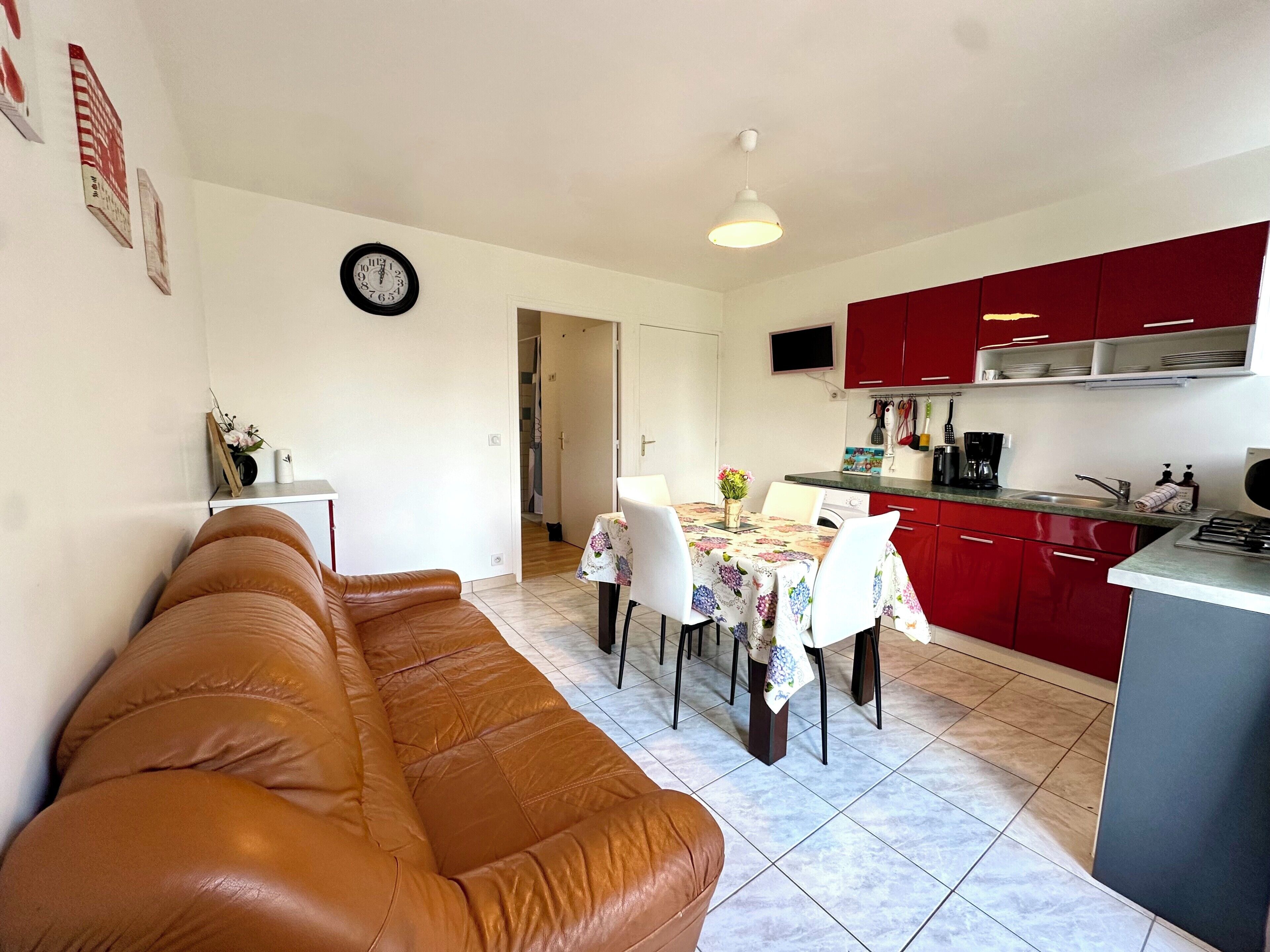 Ein Wohnzimmer mit braunem Ledersofa, einem Esstisch mit Stühlen, einer Küche mit roten Schränken und einer Uhr an der Wand.